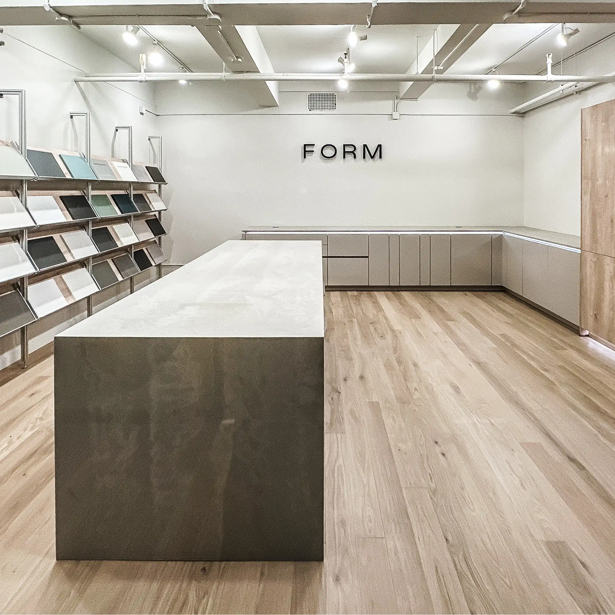 Interior of the FORM studio in New York, showcasing a minimalist and modern design. The space features a long rectangular island with a matte counter finish at the center, surrounded by light wooden flooring. One wall is equipped with shelving displaying various material samples, while the opposite side features sleek cabinetry. Overhead track lighting and the word 'FORM' illuminated on the wall accentuate the studio's contemporary aesthetic.