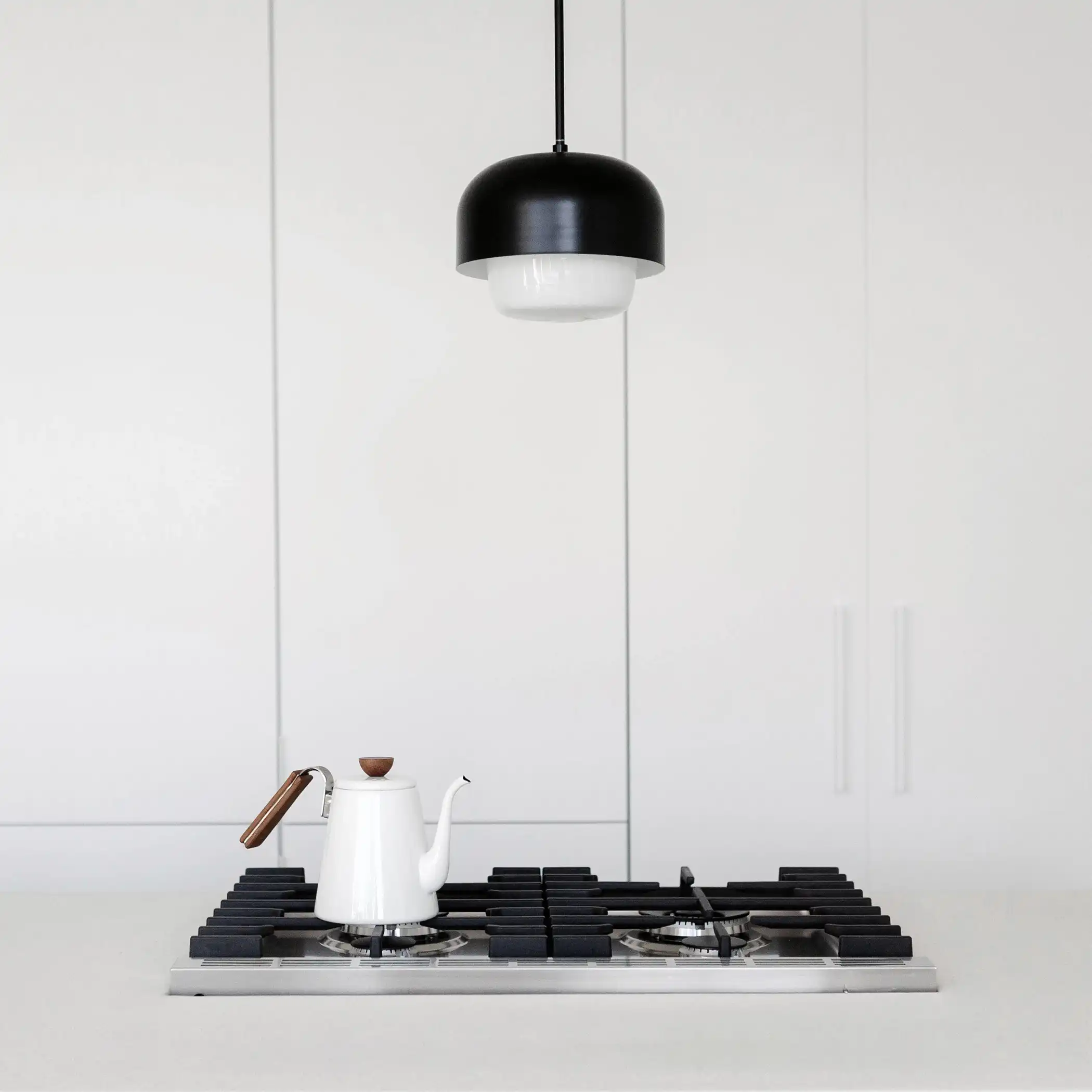 Minimalist kitchen design featuring a flush gas cooktop set into a pristine white countertop. Above the cooktop hangs a modern pendant light with a black dome and white interior, casting focused light onto the surface. A stylish white teapot with a wooden handle sits on the cooktop, adding a touch of elegance and simplicity to the scene. The background is composed of smooth white cabinetry, enhancing the clean and uncluttered aesthetic.