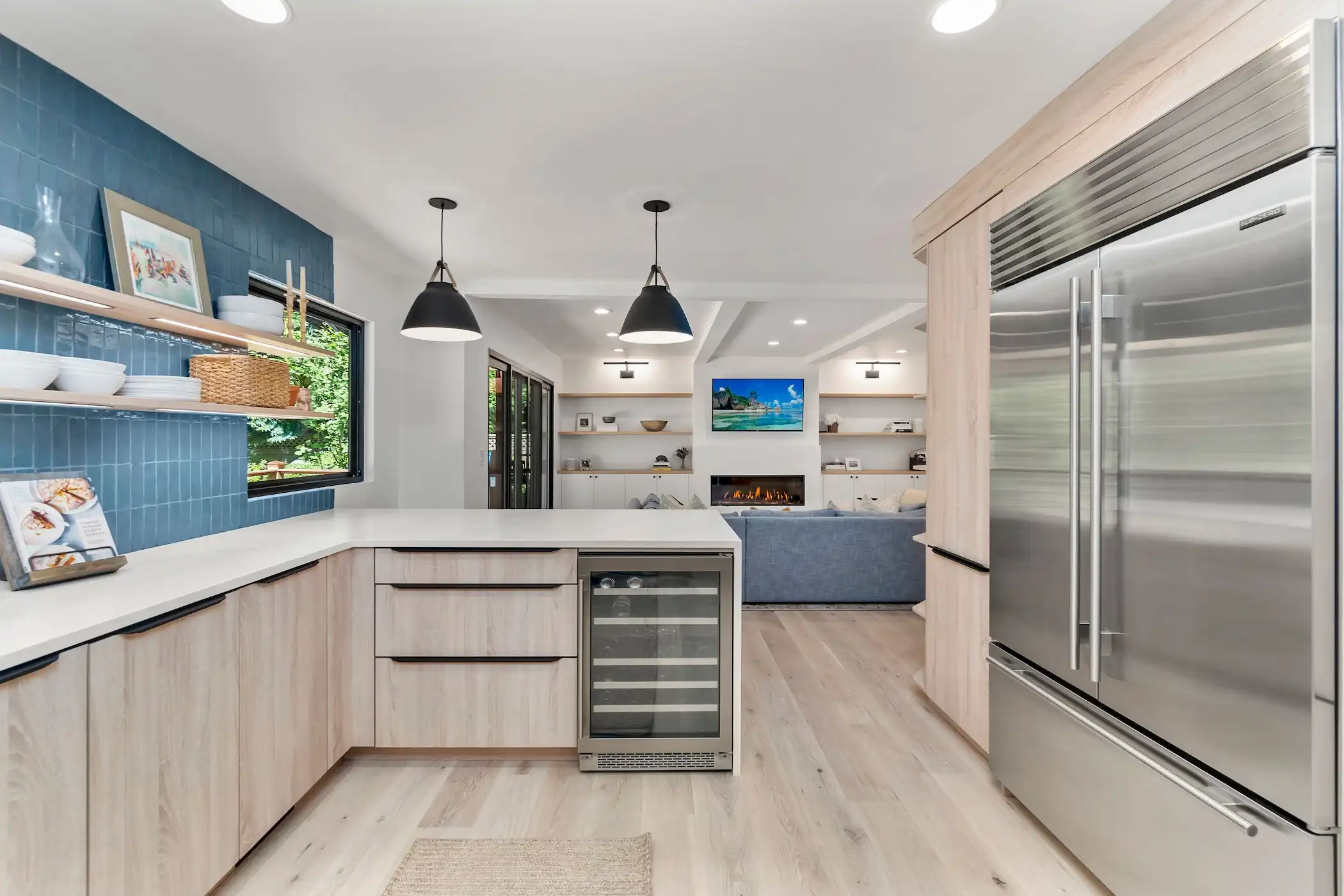 The image shows a bright and vibrant kitchen with a wood floor. The kitchen features deep blue subway tile backsplash, light wooden cabinetry, a modern stove, and white countertops. Open shelves above the counter hold dishes and cooking tools.