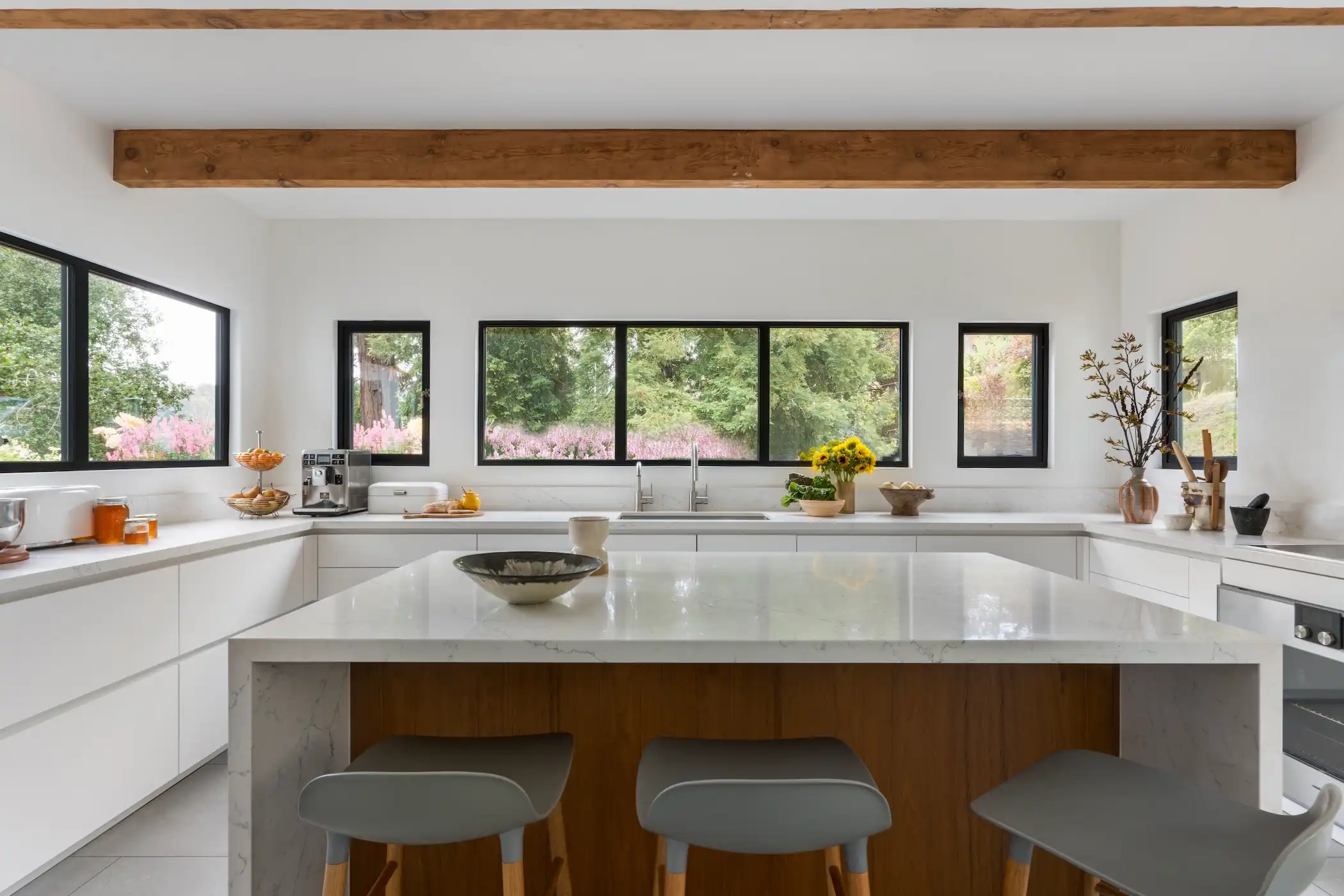 Bright and inviting kitchen featuring large windows overlooking a garden, framed by a rustic wooden beam. The space is centered around a large island with a marble surface and wood base, paired with grey stools for seating. White countertops line the perimeter, with decorative bowls and fresh flowers, contributing to the kitchen's serene and stylish atmosphere.