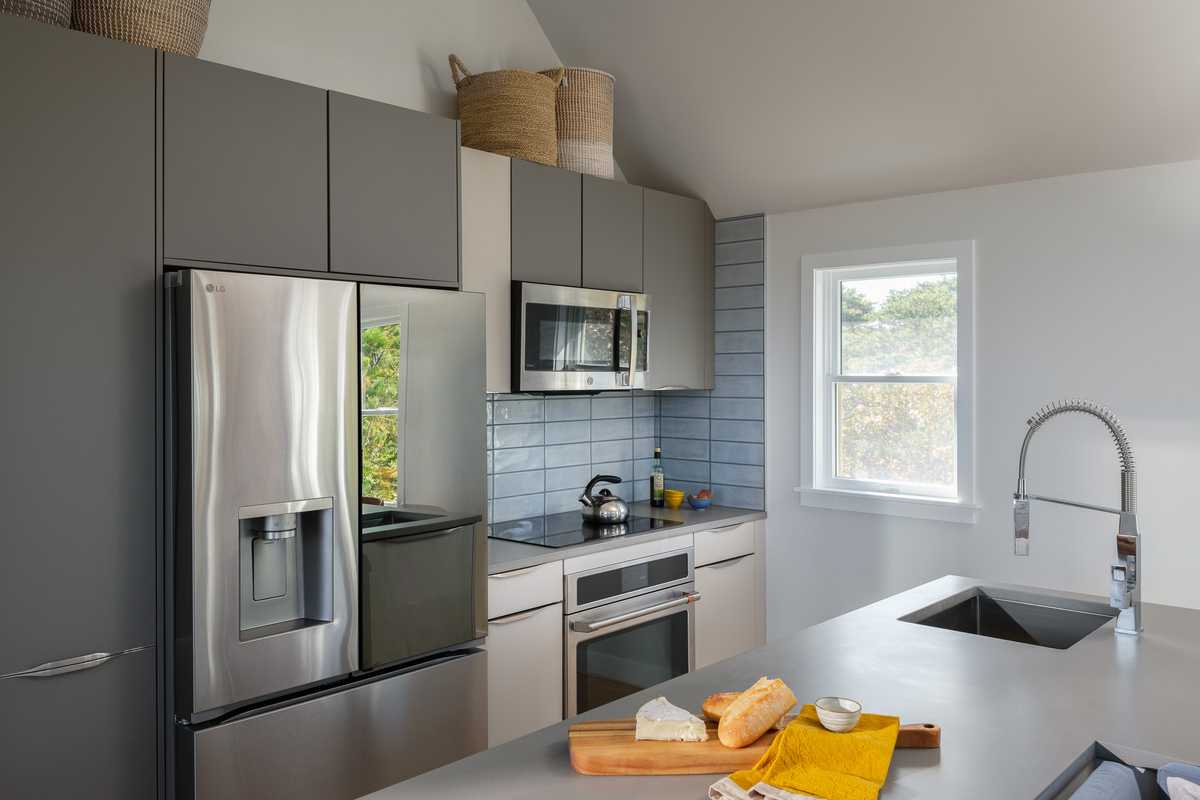 A modern kitchen with sleek Taupe Grey cabinetry, integrated handles and stainless steel appliances, including a French door refrigerator with a water dispenser and a built-in microwave above the stove. The backsplash consists of blue-gray subway tiles. A large kitchen island with a concrete countertop features an undermount sink with a high-arched spring faucet. On the countertop, a wooden cutting board holds a wedge of cheese, a sliced baguette, and a small bowl, with yellow and beige linen napkins beside it. Above the cabinets, woven baskets are used for storage. A window on the right allows natural light to enter, showing a view of trees outside.