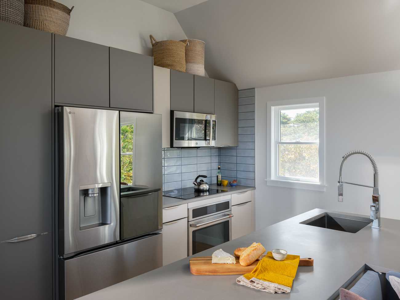 A modern kitchen with sleek Taupe Grey cabinetry, integrated handles and stainless steel appliances, including a French door refrigerator with a water dispenser and a built-in microwave above the stove. The backsplash consists of blue-gray subway tiles. A large kitchen island with a concrete countertop features an undermount sink with a high-arched spring faucet. On the countertop, a wooden cutting board holds a wedge of cheese, a sliced baguette, and a small bowl, with yellow and beige linen napkins beside it. Above the cabinets, woven baskets are used for storage. A window on the right allows natural light to enter, showing a view of trees outside.