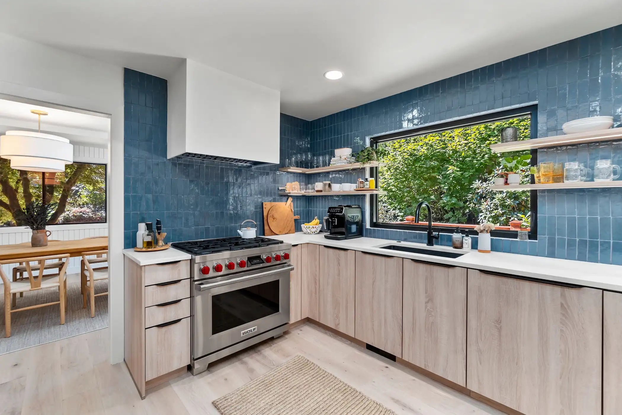 Bright and vibrant kitchen design highlighted by deep blue subway tile backsplash that reaches the ceiling, framed by a large window showcasing a lush green view. Below, light wooden cabinetry contrasts with the tiles, equipped with a modern stove and white countertops.