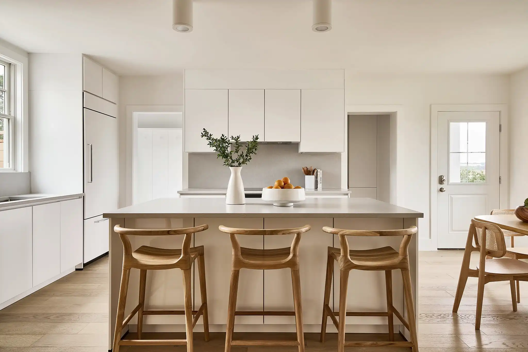 Modern kitchen design with a spacious layout, featuring white cabinetry and a large central island topped with a white countertop. The island is surrounded by elegant wooden bar stools, adding a warm contrast to the neutral tones of the room. Above the island, a minimalist vase holds greenery, and the kitchen is bathed in natural light from nearby windows, creating a welcoming and functional space.