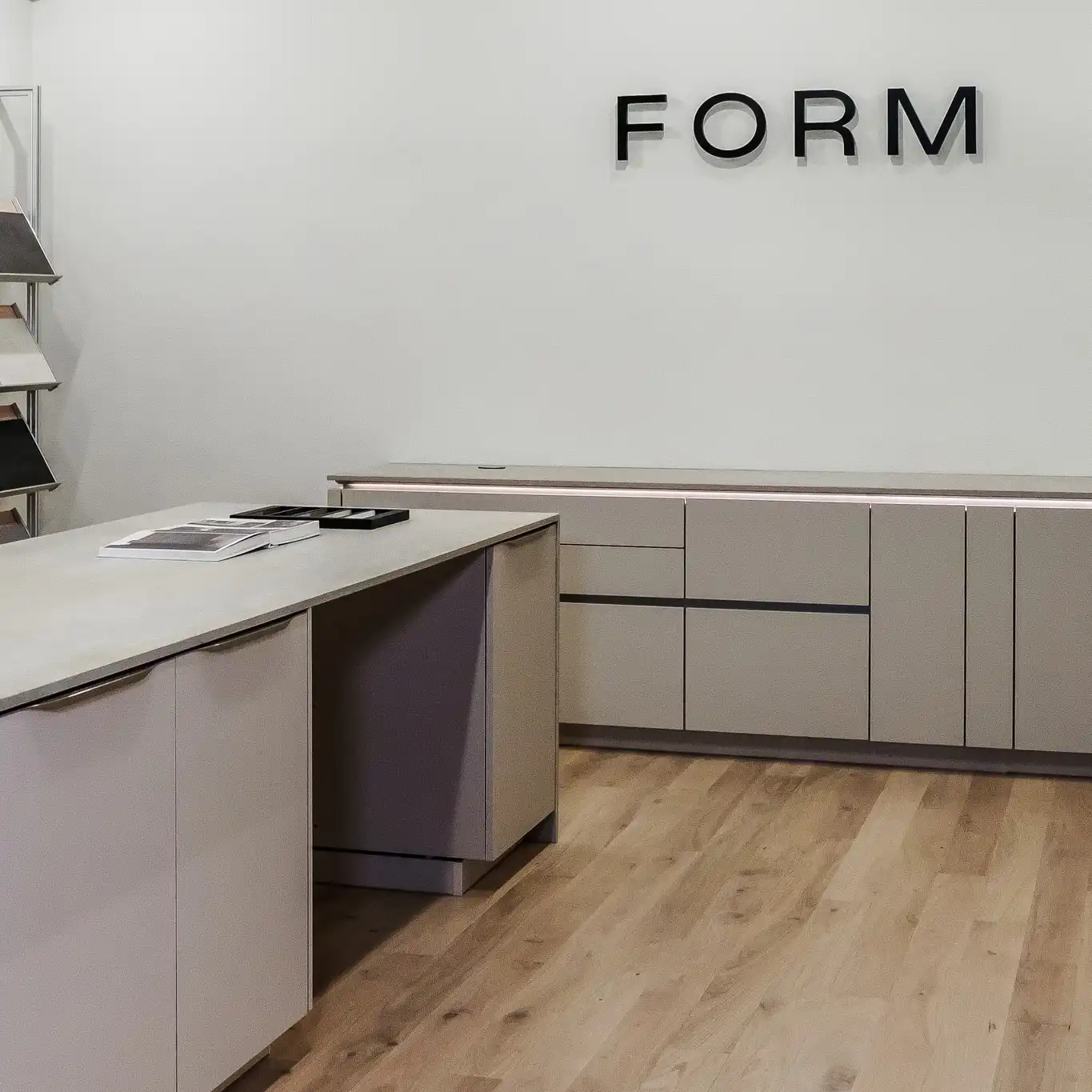 Interior view of the FORM studio in Denver, featuring a practical design workspace. The focal point is a large central island with a neutral counter and subtle cabinetry. Behind the island, a shelving unit displays an organized array of cabinet front samples ranging from tiles to wood finishes. The studio combines functionality with a clean, minimalist aesthetic, highlighted by light wooden flooring and neutral wall colors.