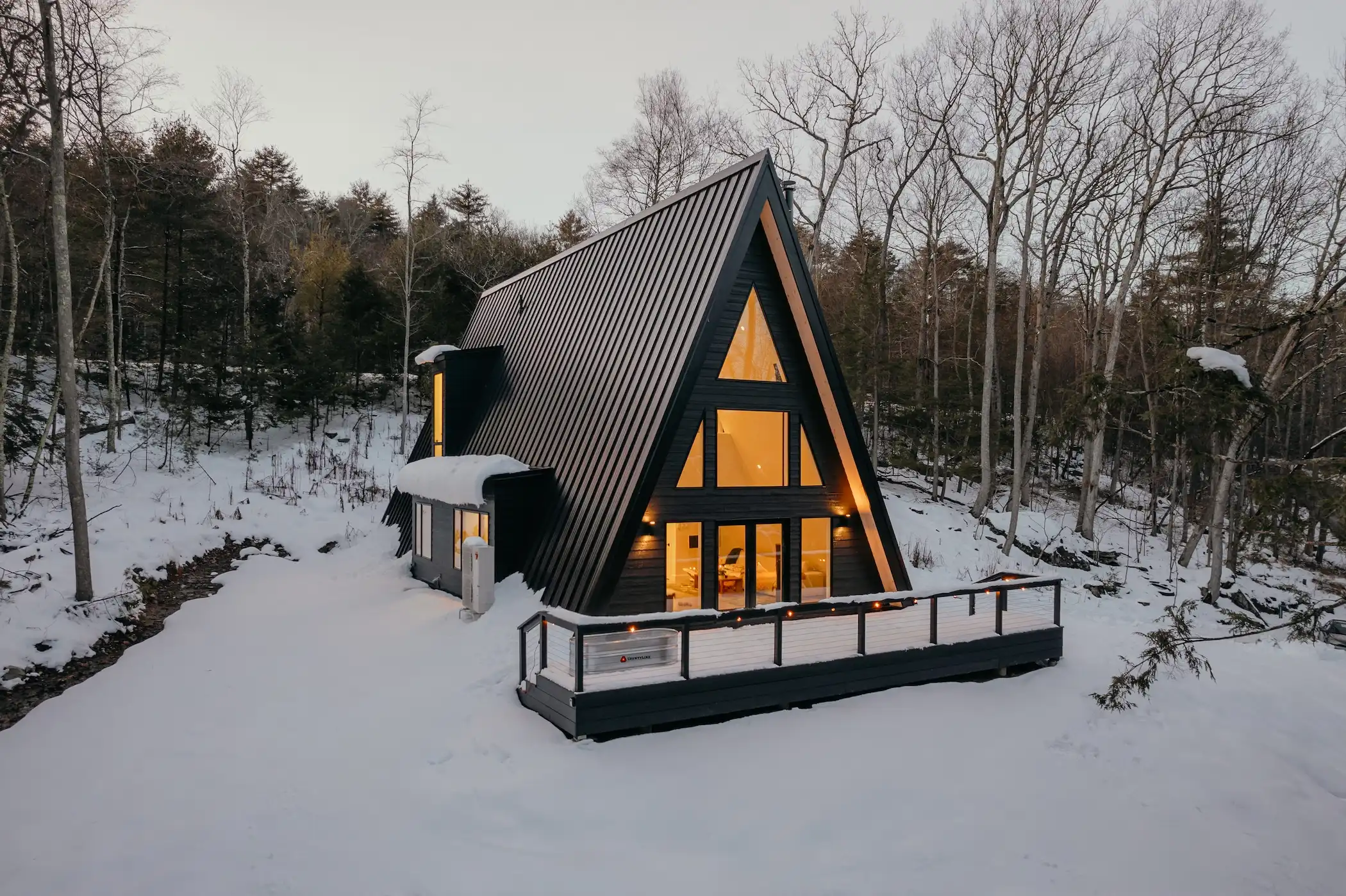 A-frame house with a roof covered in snow. It is surrounded by trees and the sky is visible in the background. The winter setting conveys a sense of freezing temperatures.