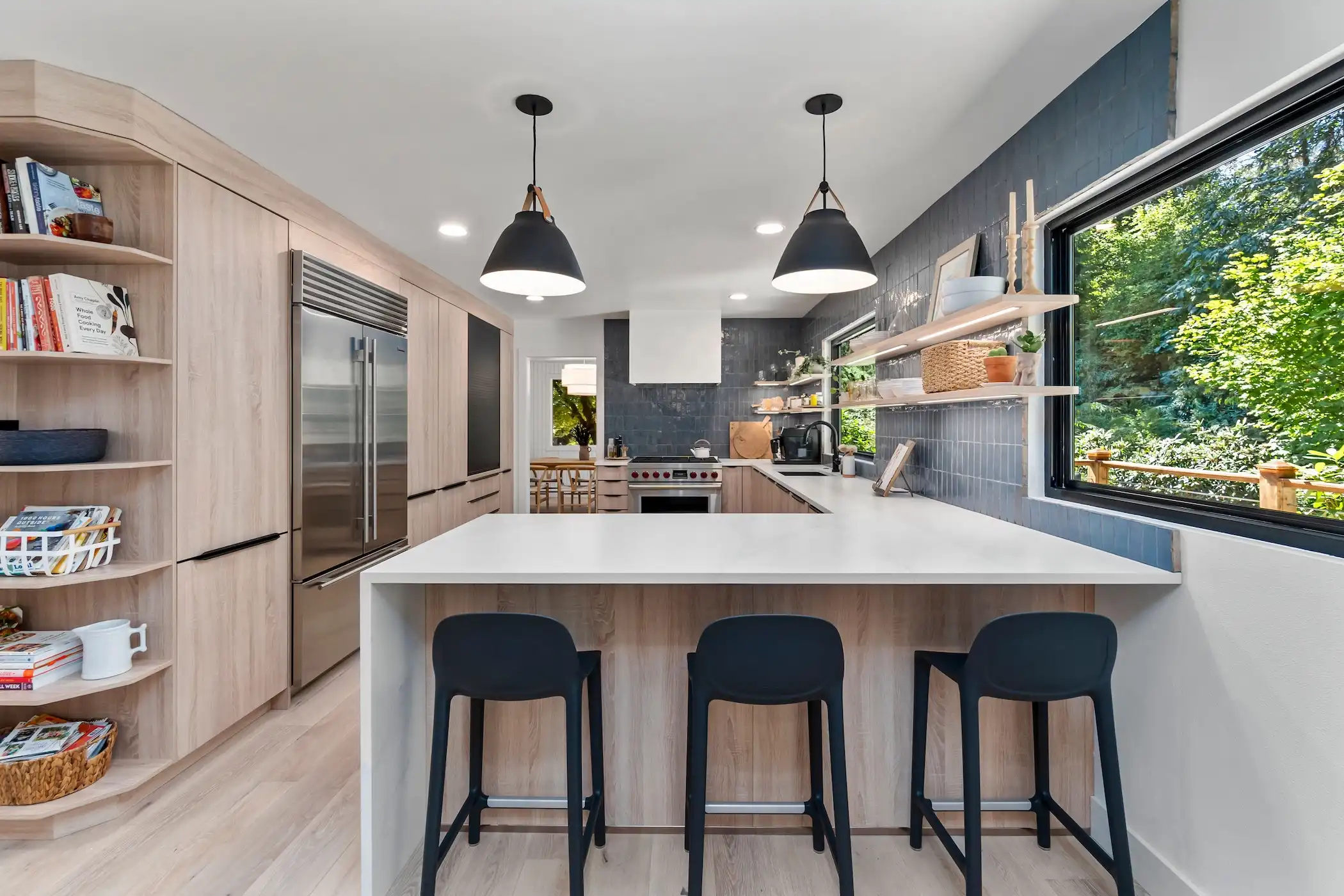 The photo is of a bright and vibrant kitchen with a table and chairs. The kitchen features deep blue subway tile backsplash reaching the ceiling, light wooden cabinetry with a modern stove and white countertops, and open shelves holding various dishes and cooking tools.