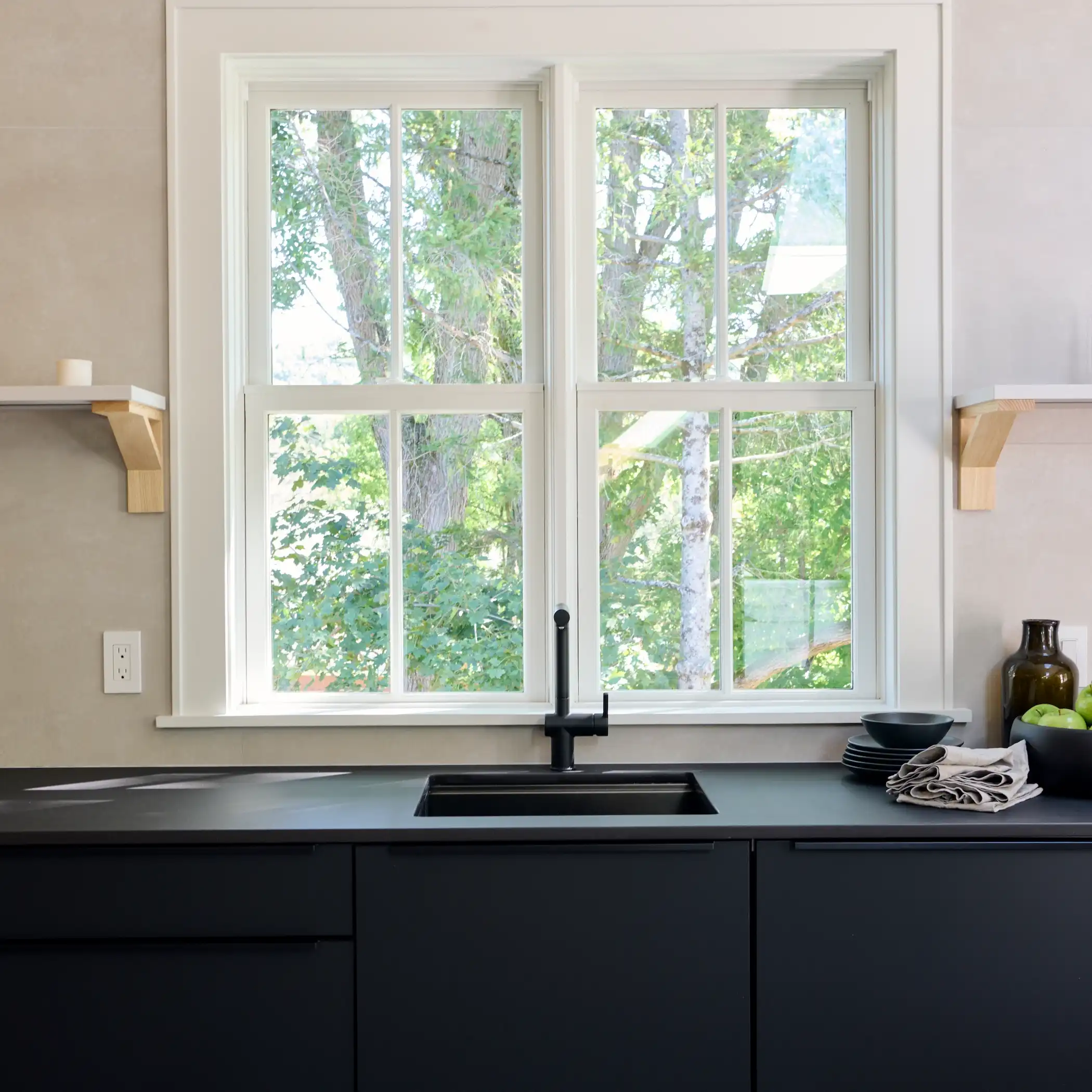 A serene kitchen setting featuring a large window that overlooks a lush green garden, framed by white millwork. Below the window is a sleek black countertop with a modern black sink and faucet.