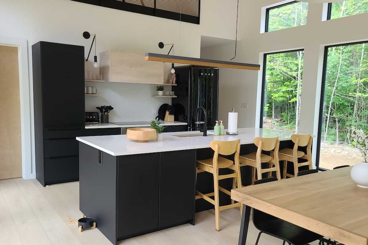 Modern, open-concept kitchen, large windows, and a sleek, black-and-oak kitchen island. The space features two large, black pendant lights and a black Smeg refrigerator, framed by natural wood accents and surrounded by ample natural light from the surrounding forest.