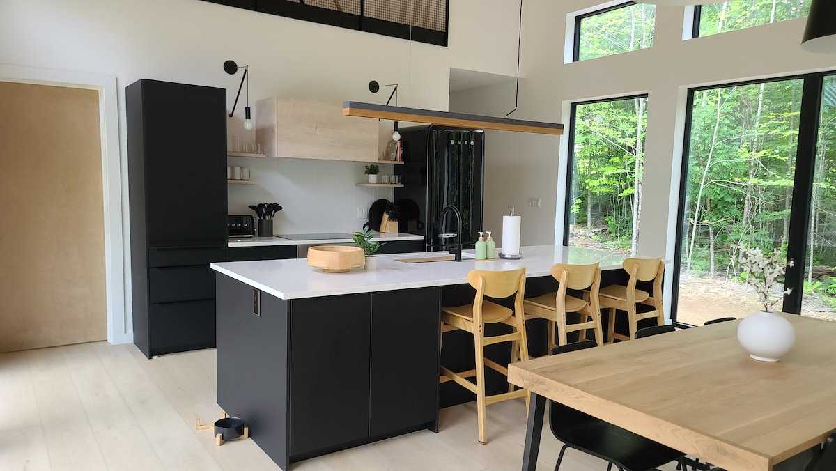 Modern, open-concept kitchen, large windows, and a sleek, black-and-oak kitchen island. The space features two large, black pendant lights and a black Smeg refrigerator, framed by natural wood accents and surrounded by ample natural light from the surrounding forest.