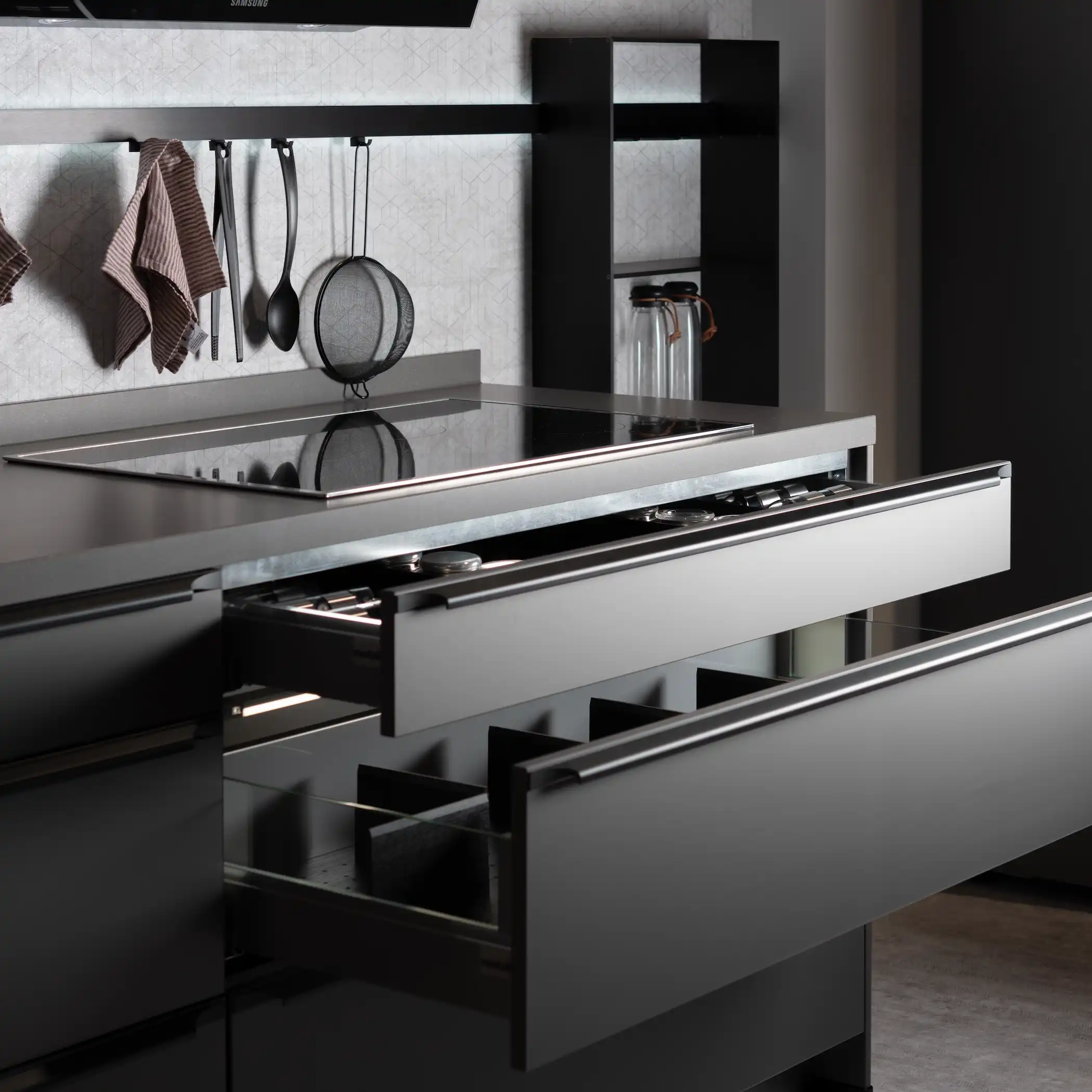 A sophisticated graphite kitchen workspace featuring a sleek, modern drawer system with a dark countertop. The drawer is open, displaying organized compartments for utensils alongside a high-end induction cooktop. Above the cooktop, kitchen tools hang neatly on a wall-mounted rack against a textured backdrop, emphasizing functionality and style in kitchen design