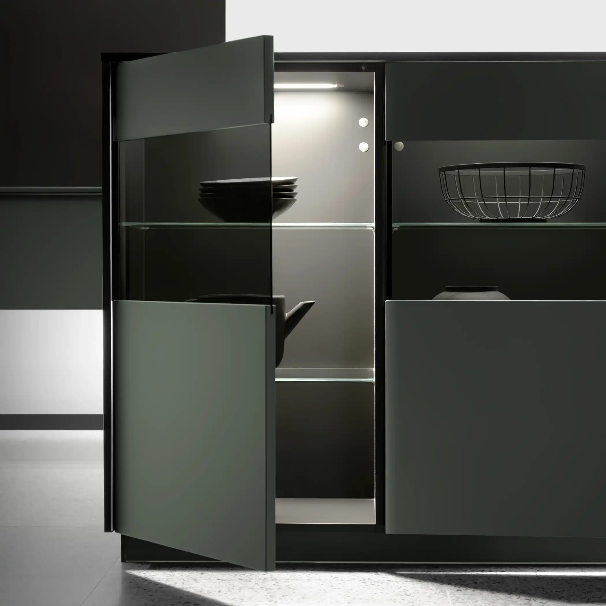 A close-up view of a modern, high-end kitchen cabinet in a deep green finish, partially open to reveal neatly arranged shelves. The cabinet features an illuminated interior showcasing stacked plates, various kitchen tools, and a basket. The sleek design and subtle lighting highlight the cabinet’s sophisticated look and functionality.