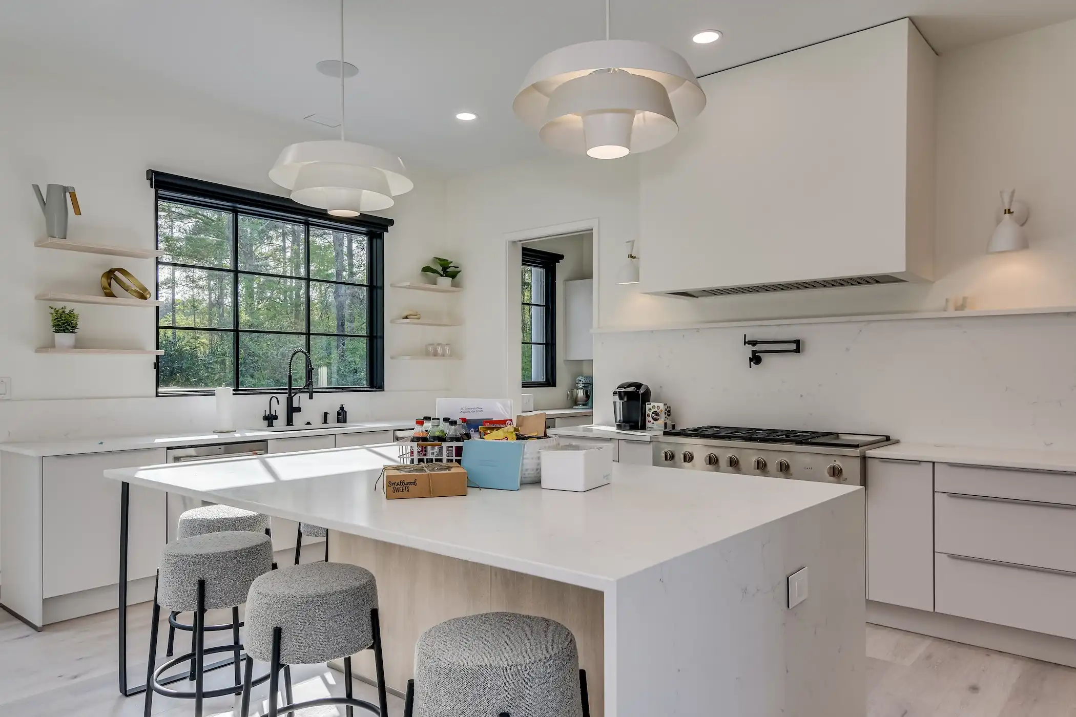 Bright and inviting kitchen featuring large windows overlooking a garden, framed by a rustic wooden beam. The space is centered around a large island with a marble-like surface and wood base, paired with grey stools for seating. White countertops line the perimeter, adorned with decorative bowls and fresh flowers, contributing to the kitchen's serene and stylish atmosphere.