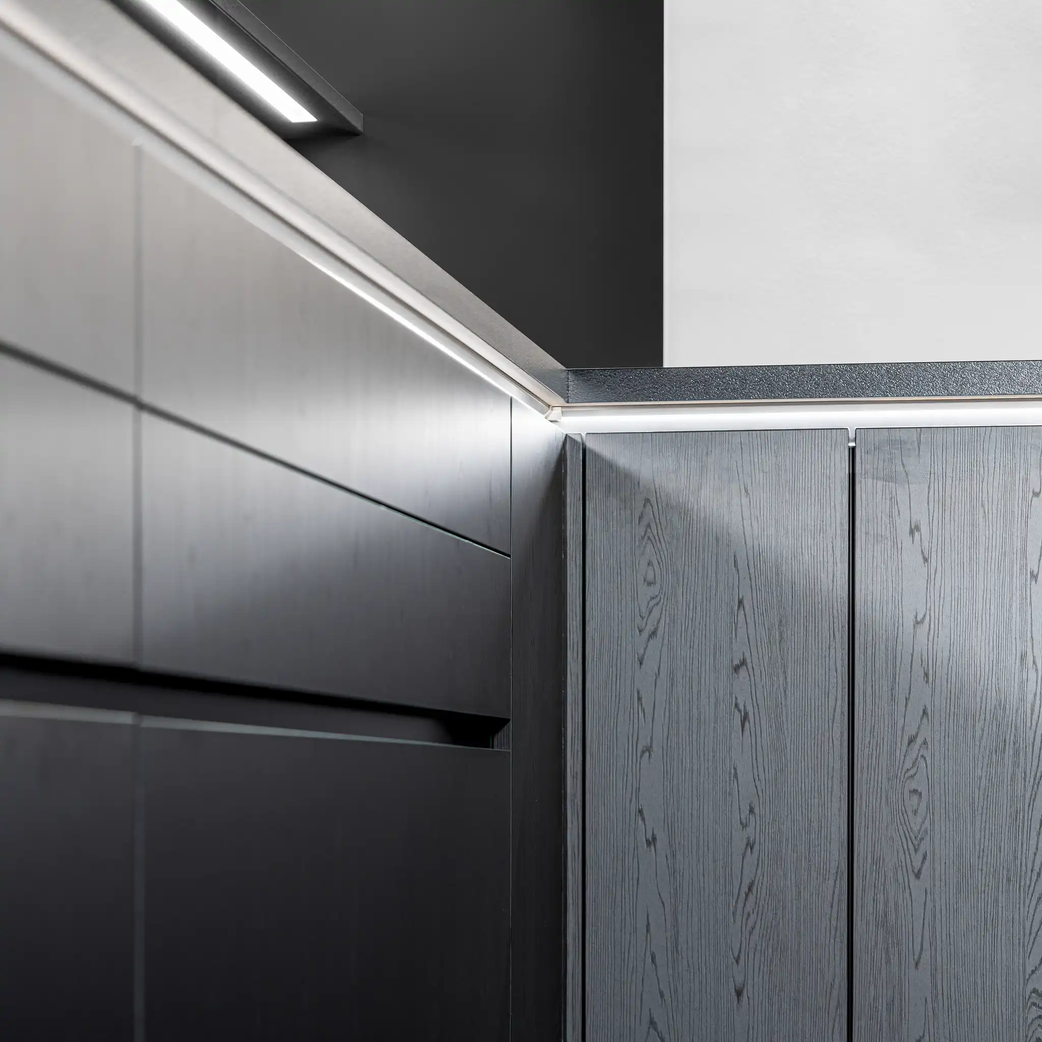 Close-up view of a modern kitchen's corner, showcasing textured Nero Oak Reproduction cabinetry with subtle wood grain patterns. The cabinets feature a sleek, handle-less design, emphasizing a minimalist aesthetic. Under-counter lighting casts a soft glow, highlighting the unique texture and enhancing the contemporary feel of the kitchen design.