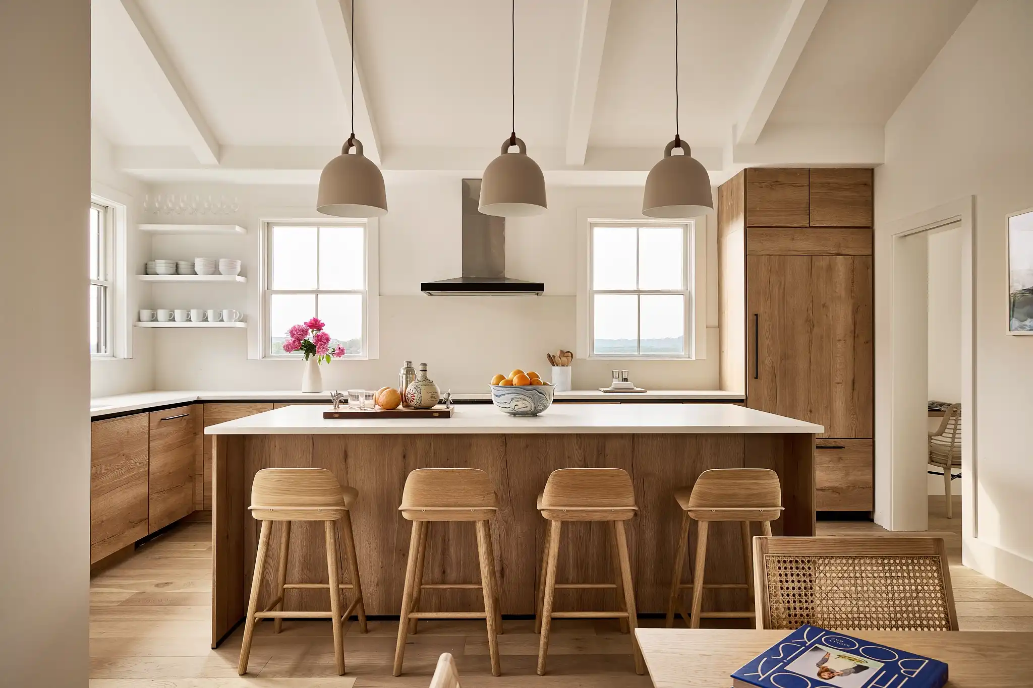 Cozy and inviting kitchen space featuring natural wood cabinetry and a large central island with a white countertop. The island is surrounded by wooden bar stools, creating a welcoming area for socializing. Above, three muted gray pendant lights provide soft illumination. The kitchen is accented with open shelving displaying ceramic dishes, and fresh flowers add a splash of color. Large windows let in ample natural light, enhancing the warm, earthy tones of the room.