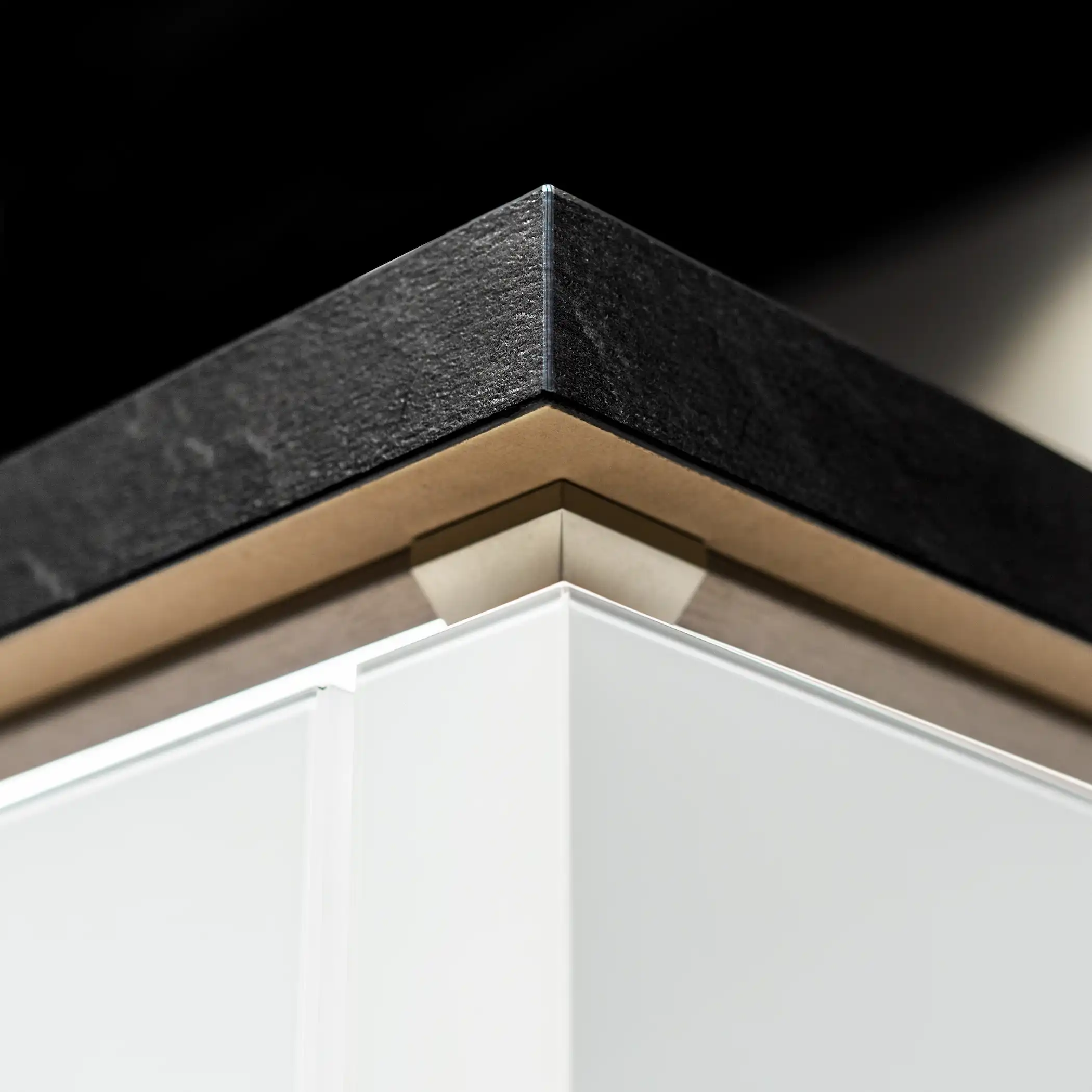 Close-up view of the top corner of a modern kitchen cabinet, featuring a layered design with a textured black top, a thin metallic trim, and a white cabinet face. The contrasting textures and colors highlight the cabinet’s contemporary styling, showcasing meticulous attention to detail and a sophisticated material palette.