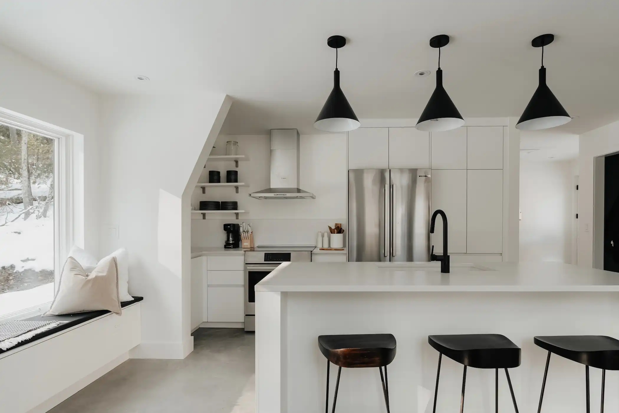 Modern and minimalist kitchen design with clean white cabinetry and an island with a white countertop and contrasting black faucet. Above the island, three black pendant lights add a contemporary touch against the bright backdrop. The kitchen includes black bar stools and features built-in shelves stocked with kitchen essentials. A window seat with a cushion provides a cozy nook beside a snowy outdoor view, blending comfort with functional design.