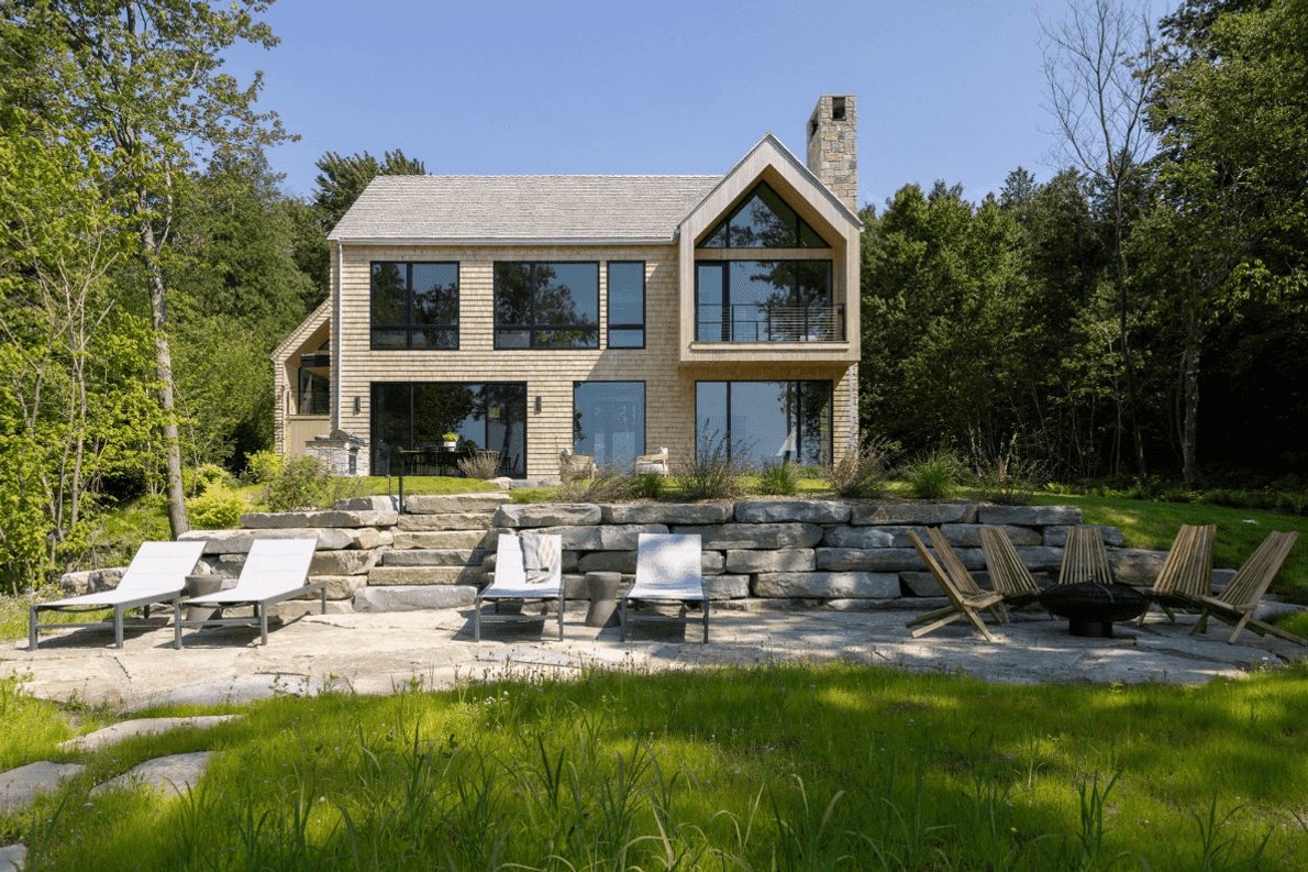 Modern lakefront home with a two-story design featuring large glass windows that provide expansive views and connect the interior to the surrounding nature. The exterior showcases cedar wood siding and a gabled roofline, blending seamlessly with the lush forested backdrop. A tiered stone patio includes lounge chairs and seating arranged around a fire pit, offering a relaxing outdoor space. Stone steps lead up to the home, enhancing the rustic yet refined aesthetic of this peaceful retreat.