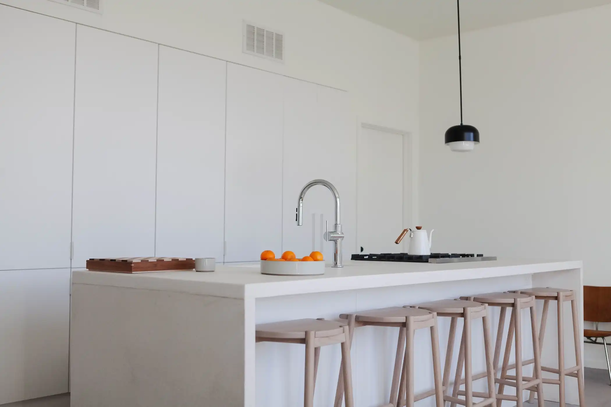 Minimalist kitchen design with a spacious white island, featuring bar seating and light wooden stools. The clean, white cabinetry blends seamlessly with the walls, creating a unified and uncluttered look. A single black pendant light hangs above, providing a modern contrast. The island is equipped with a sleek sink and cooktop, with a few oranges adding a pop of color to the neutral palette.