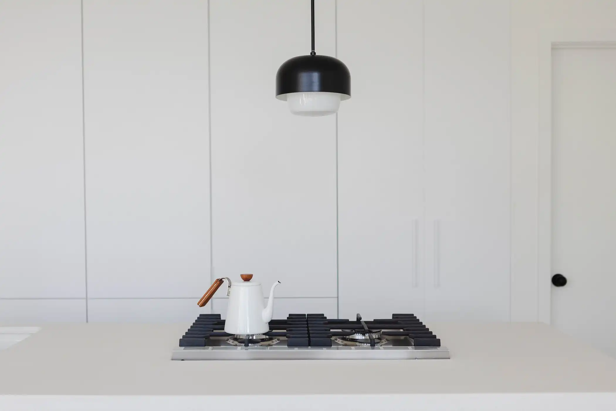 A minimalist white kitchen with a spacious island featuring bar seating and light wooden stools. The clean white cabinetry blends seamlessly with the walls, creating a unified and uncluttered look. A black pendant light provides a modern contrast, and the island is equipped with a sink and cooktop, with oranges adding a pop of color to the neutral palette.