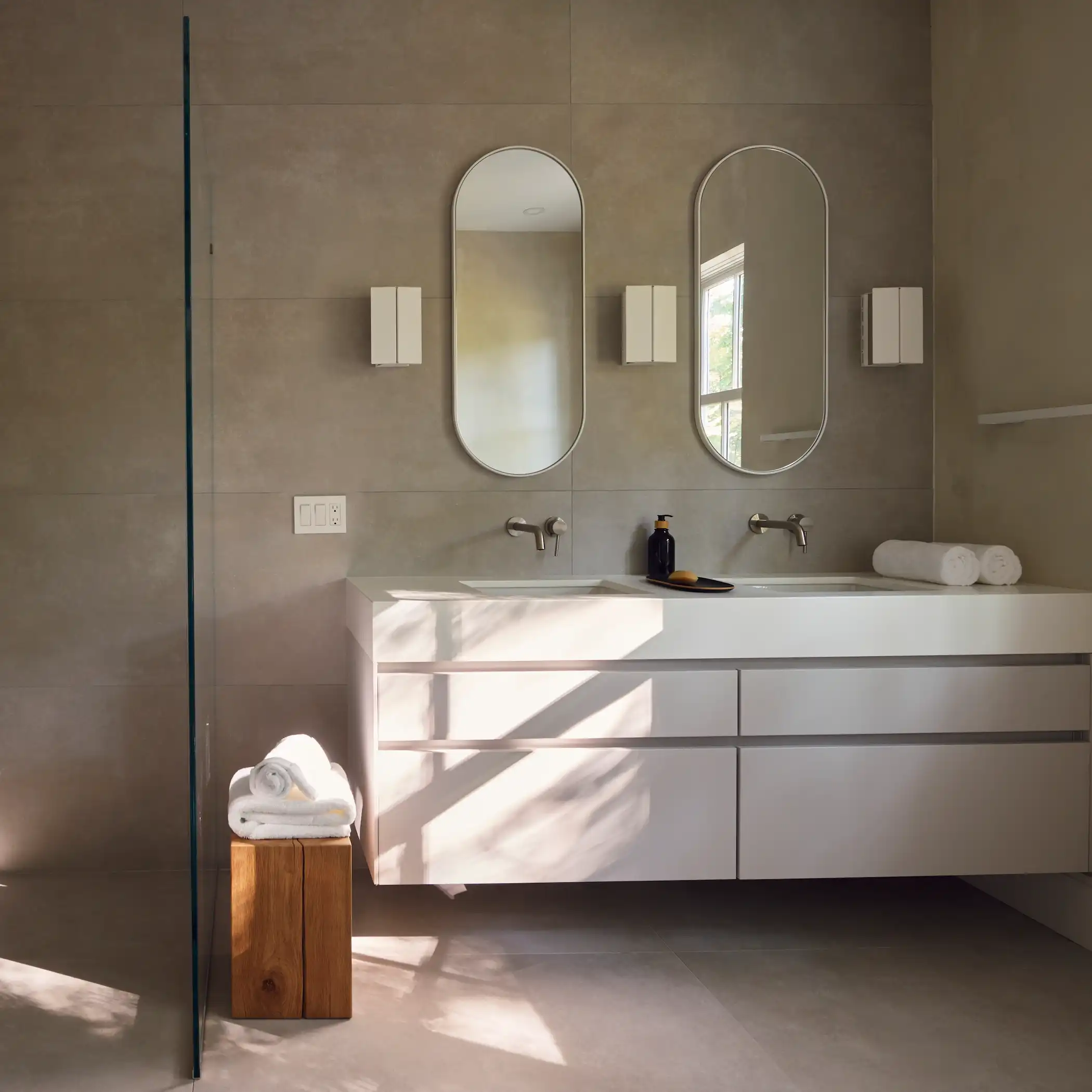 This minimalist bathroom design features a serene, monochromatic color palette with concrete-like walls and floor. A floating white vanity is fitted with dual sinks and features a clean, sleek design. Above the vanity, two oval mirrors are complemented by simple, modern lighting fixtures. Natural light filters through a window, casting intriguing shadows across the space, enhancing the tranquil and refined aesthetic. A small wooden stool adds a touch of warmth to the otherwise cool-toned setting.