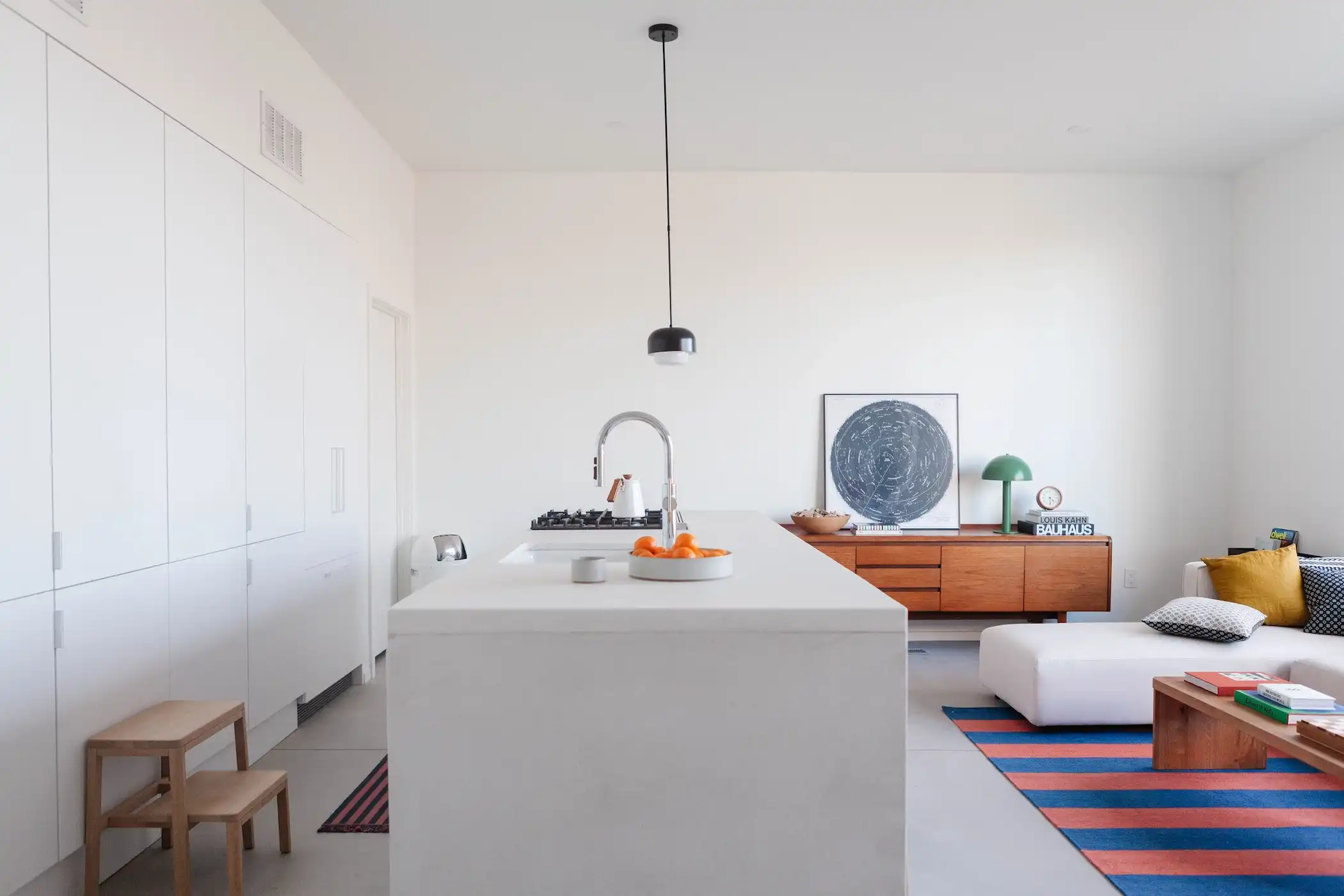 A minimalist white kitchen with a sink and a sleek island, featuring bar seating and light wooden stools. The clean, white cabinetry blends with the walls, creating a unified look. A black pendant light hangs above, providing a modern contrast. Oranges add a pop of color to the neutral palette.