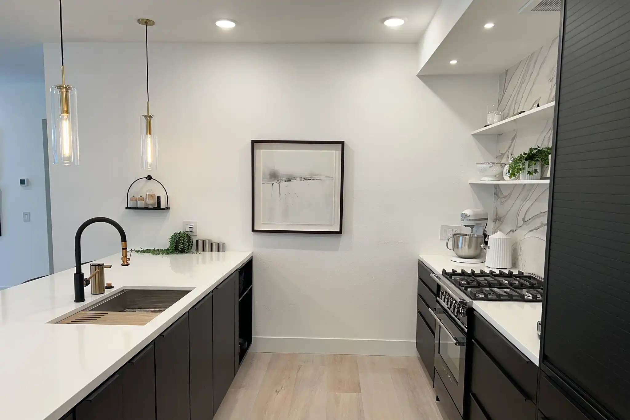 Modern kitchen design featuring sleek black cabinetry and a white countertop. A central sink with a high, black faucet complements the aesthetic, illuminated by glass pendant lights. To the right, a gas range is set against a porcelain backsplash, enhancing the luxurious feel. Floating shelves with greenery and a framed artwork add a touch of elegance and personality to the space.