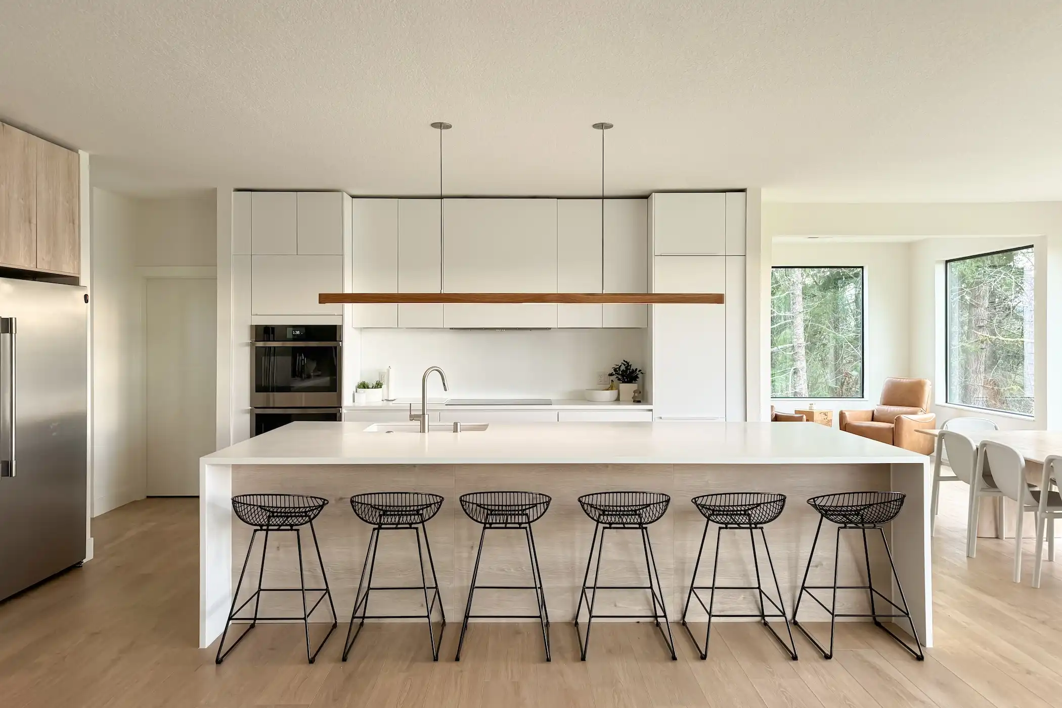 Contemporary kitchen bathed in natural light, showcasing a large white island with sleek bar stools and a harmonious blend of white cabinetry and our Oak Montreal cabinets. The space is well-equipped with high-end appliances and minimalistic pendant lighting. An inviting nook with a comfortable chair next to the window creates a serene spot for relaxation, all set against a backdrop of lush greenery visible through large windows.