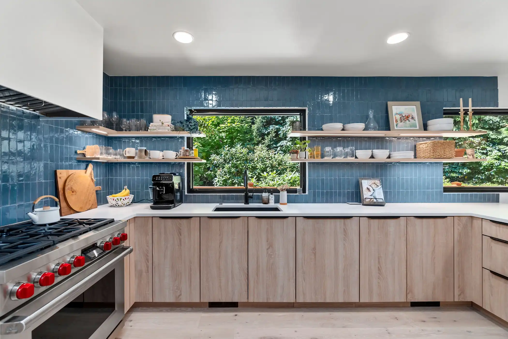 Bright and vibrant kitchen design highlighted by deep blue subway tile backsplash that reaches the ceiling, framed by a large window showcasing a lush green view. Below, light wooden cabinetry contrasts with the tiles, equipped with a modern stove and white countertops. Open shelves above the counter hold a variety of dishes, cooking tools, and decorative items, adding functionality and charm to the space.